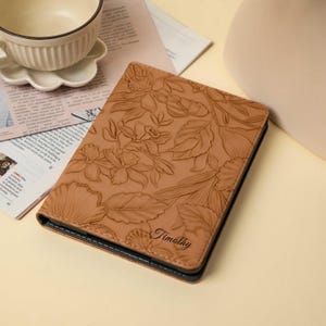May include: A tan leather journal with a floral embossed design. The journal has a black border and the name "Timothy" is printed on the lower right corner. A white teacup and saucer sit on a newspaper in the background.