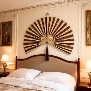 May include: A large decorative fan with brown and beige panels is mounted on a wall above a bed. The fan has a dark brown tassel. The bed has white pillows and a beige headboard. Two lamps flank the bed. The room has a classic design.