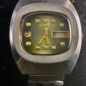 Op de afbeelding: Vintage Rado polshorloge met een zilverkleurige kast en band. De wijzerplaat is groen met goudkleurige wijzers en markeringen. De datumweergave toont "1 MON 6". Het horloge heeft een rechthoekige wijzerplaat en een roestvrijstalen band.