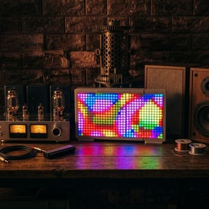 Op de afbeelding: Een rechthoekig LED-pixeldisplay met een levendig, veelkleurig abstract patroon van vierkantjes. Het apparaat staat op een houten oppervlak naast een zilverkleurige buizenversterker met twee analoge meters en een houten luidsprekerkast.