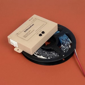 Könnte beinhalten: Ein beigefarbenes Steuergerät mit der Aufschrift "Elmaster" und einem Schalter liegt auf einer schwarzen Rolle mit LED-Streifen. Die Rolle ist teilweise abgerollt und zeigt blaue und weiße Lichter. Drähte sind sichtbar.