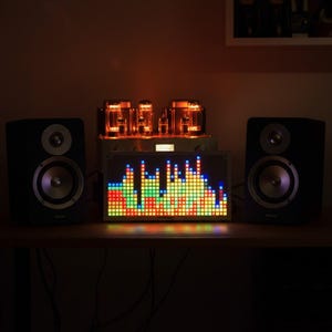 Op de afbeelding: Een paar zwarte boekenplankluidsprekers geflankeerd door een zilveren buizenversterker en een rechthoekig LED-muziekvisualisatiedisplay. De visualizer toont een kleurrijk raster van rode, gele, groene en blauwe vierkanten.