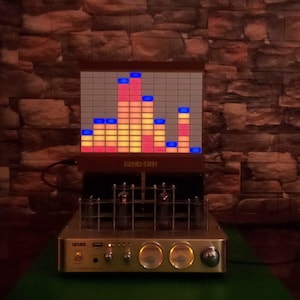 May include: A vacuum tube audio amplifier with a metallic gold front panel featuring two circular analog gauges, control knobs, and a power switch. Above the tubes, a rectangular display shows a glowing red, orange, and blue audio spectrum.