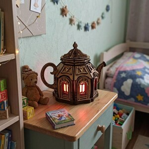 Puede incluir: Una lámpara de madera cortada con láser en forma de tetera con un diseño arquitectónico intrincado, que presenta ventanas arqueadas y una tapa abovedada. La lámpara emite una cálida luz naranja desde el interior, sobre una superficie de madera.