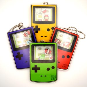 Puede incluir: Cinco consolas de juegos portátiles de tamaño llavero de estilo Game Boy con pantallas y botones de diferentes colores. Las consolas están dispuestas en círculo, con la verde en el centro.