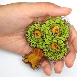 May include: A green and brown tree-shaped acrylic charm with three golden stars. The charm is held in a hand.