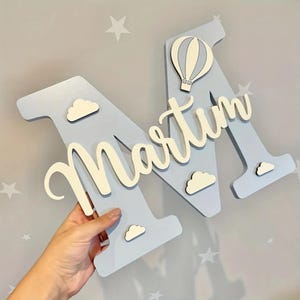 Peut inclure: Une lettre "M" en bois bleu clair avec le nom "Martim" en cursive, décorée de nuages blancs et d'une montgolfière. La lettre est tenue par une main contre un mur gris avec des motifs d'étoiles.