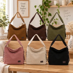 Puede incluir: Seis bolsos de hombro estilo hobo hechos de tela de pana, dispuestos en dos filas. Los colores incluyen marrón claro, marrón oscuro, verde oliva, rosa empolvado, crema y negro. Cada bolso cuenta con una correa ancha y una etiqueta de marca.