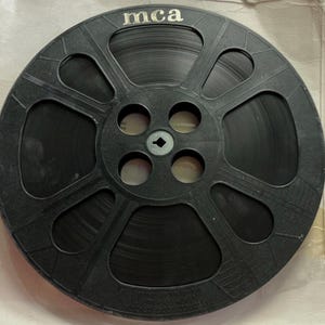 May include: A black plastic film reel featuring the text mca printed at the top. The circular reel has a central hub with four circular cutouts and eight larger trapezoidal openings around the perimeter, holding a roll of dark film.