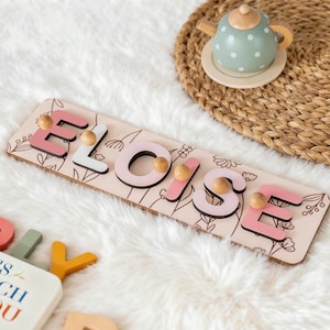 May include: A wooden name puzzle spelling "Eloise" in pink and white letters with wooden pegs. The puzzle sits on a light beige board with floral line art. A small toy teapot and a woven mat are in the background.
