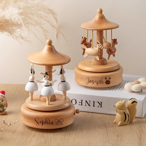 May include: Two wooden music boxes with a carousel theme. One features ballerina figurines, the other has horse figurines. Both are light brown with a domed top and a circular base. The ballerina music box has the name "Sophia" engraved on it.