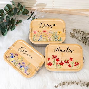 Op de afbeelding: Drie rechthoekige bamboe lunchboxen met bloemmotieven en de namen "Daisy", "Olivia" en "Amelia". Elke doos heeft een ander aquarel bloemarrangement in roze, rood, geel en blauw. De namen zijn in elegant zwart schrift.