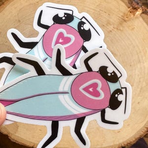 May include: Two blue and pink cartoon cockroaches with hearts on their backs. The cockroaches are on a wood background.