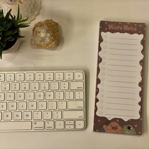 May include: A white keyboard with a brown and gold notepad that says "Micro Notes" on it. The notepad has a white lined area with a dotted border and a cartoon animal illustration at the bottom. A blue pen is on the right side of the notepad.