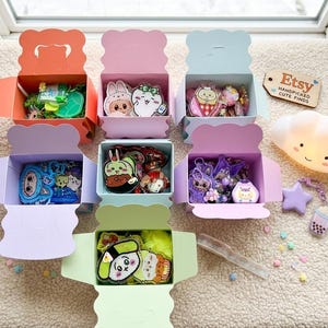 May include: Seven open pastel-colored cardboard boxes filled with various acrylic charms and keychains. A wooden tag reads Etsy Handpicked Cute Finds. A small white cloud-shaped lamp, a purple star charm, and a boba tea charm sit nearby.