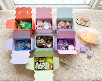 May include: Seven open pastel-colored cardboard boxes filled with various acrylic character charms and keychains. A wooden tag reads Etsy HANDPICKED CUTE FINDS. A white cloud-shaped lamp, a purple star charm, and a boba tea charm sit nearby.