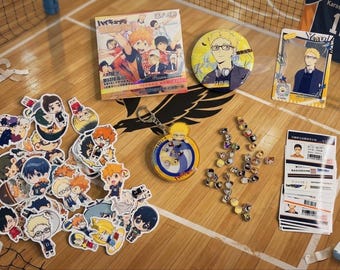 Op de afbeelding: Een verzameling Haikyuu anime merchandise, waaronder een vierkant boekje, een ronde speld, een rechthoekige kaart, een shaker sleutelhanger, een stapel karakterstickers, kleine metalen pins en een stapel identificatiekaarten met barcodes.