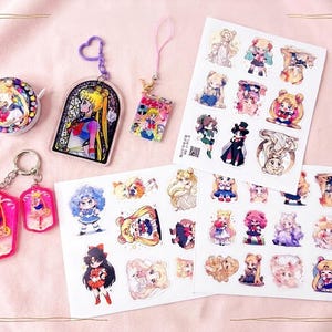 Op de afbeelding: Een verzameling Sailor Moon merchandise, waaronder twee acryl sleutelhangers met hart- en lusbevestigingen, een ronde pin, een kleine rechthoekige bedel en twee stickervellen met diverse chibi-stijl karakterillustraties.