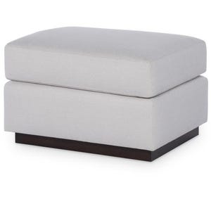 Puede incluir: Un otomán tapizado de color gris claro con una base marrón oscuro. El otomán tiene forma rectangular con un diseño de dos niveles. El cojín superior es ligeramente más grande que la base.