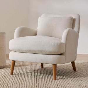 May include: Cream-coloured armchair with a matching cushion. The chair features a rounded back and armrests, supported by wooden legs. It is positioned on a textured rug. The design is simple and modern.