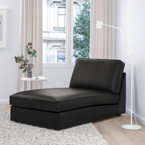 May include: A black leather chaise lounge with a rectangular base and an angled backrest. The seat and back have stitched detailing. The chaise is placed on a white rug in front of a window with white curtains.