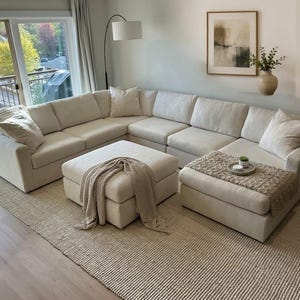 May include: A large, off-white sectional sofa with a matching square ottoman and several throw pillows. A textured beige throw blanket is draped over the ottoman, and a patterned runner with a small decorative tray sits on the chaise section.