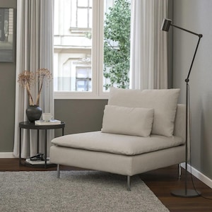 May include: A beige armchair with a matching lumbar pillow and a large back cushion. The chair has silver legs and sits on a gray rug. A black side table with a vase of dried flowers and a floor lamp are nearby.