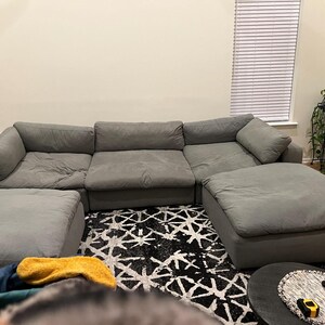 May include: A large, modular gray sectional sofa with two matching ottomans. The sofa is arranged in a U-shape, with plush cushions. The room features a patterned rug and a plant stand.