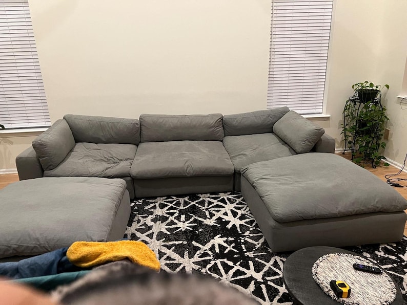 May include: A large, modular gray sectional sofa with two matching ottomans. The sofa is arranged in a U-shape, with plush cushions. The room features a patterned rug and a plant stand.