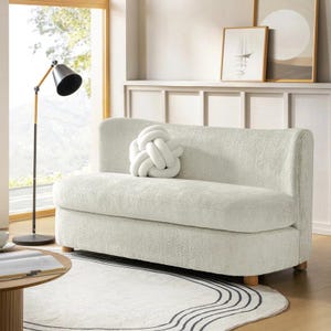 May include: A cream-colored, two-seater sofa with a textured fabric and wooden legs. A white knot-shaped pillow rests on the seat. The sofa sits on a white rug with black wavy lines. A black and gold floor lamp stands nearby.