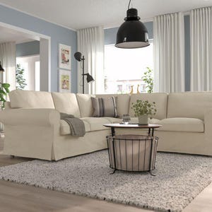 Puede incluir: Un gran sofá seccional color crema con brazos enrollados y una base con falda. Una pequeña mesa de centro redonda con una tapa de madera oscura y una base de cesta de metal se encuentra sobre una alfombra gris claro. La habitación tiene cortinas blancas y una lámpara colgante negra.