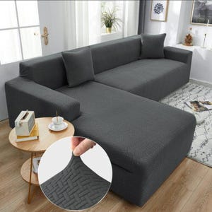 May include: Dark gray L-shaped sofa cover with a textured pattern. The cover includes two matching throw pillows. A close-up shows the fabric's stretch and weave. The sofa is in a well-lit room with a small side table.