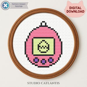 May include: A cross-stitch design of a pink handheld digital pet game, framed in a wooden hoop. The game's screen is light green with a pixelated character. The design includes the text "DIGITAL DOWNLOAD" and "STUDIO CATLANTIS".