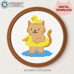 May include: A cross stitch pattern featuring a light brown cat wearing a yellow duck-shaped swim ring, with a small yellow duck on its head, standing in a blue puddle. The design is framed in a wooden hoop with text reading Pattern Keeper TESTED.