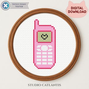 Può includere: Un motivo a punto croce raffigurante un telefono cellulare rosa con uno schermo verde chiaro che mostra un'icona a forma di cuore nero. Il disegno è incorniciato in un telaio circolare in legno. Il testo include Pattern Keeper TESTED, DIGITAL DOWNLOAD e STUDIO CATLANTIS.