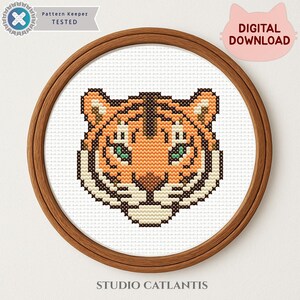 Puede incluir: Un dise&ntilde;o de punto de cruz de una cabeza de tigre en un aro de bordado de madera. El tigre tiene pelaje naranja y marr&oacute;n con ojos verdes. Se ve el texto "Pattern Keeper TESTED" y "DIGITAL DOWNLOAD".