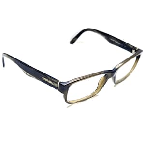 Può includere: Occhiali Prada con montatura rettangolare. Gli occhiali presentano una combinazione di colori grigio scuro e verde oliva. Il logo Prada è visibile sulla stanghetta sinistra degli occhiali.
