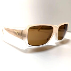 May include: Cream-colored Prada sunglasses with brown lenses. The rectangular frames have a subtle, translucent quality. The Prada logo is visible on the temples. A stylish accessory for eye protection.