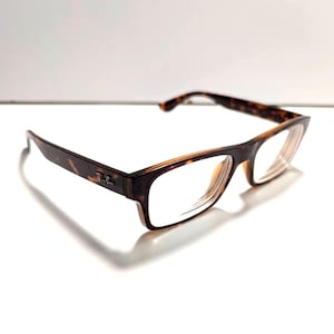 May include: A pair of Ray-Ban eyeglasses with a tortoiseshell pattern on the frames. The glasses have a rectangular shape with clear lenses. The Ray-Ban logo is visible on the left arm of the glasses.