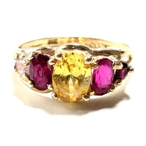 Puede incluir: Un anillo de oro con una gran piedra preciosa amarilla ovalada flanqueada por dos piedras preciosas magenta ovaladas y piedras más pequeñas rosas y magenta. El anillo tiene un diseño clásico y un acabado pulido.