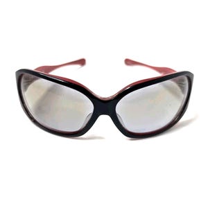 May include: Black and red-framed sunglasses with a cat-eye shape. The lenses are a light silver color. The arms of the sunglasses are red, and the frame is black. The sunglasses are set against a white background.