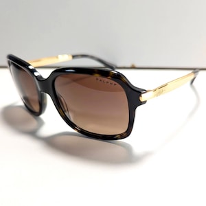 Könnte beinhalten: Quadratische Sonnenbrille mit Schildpattmuster auf den Rahmen und braunen Verlaufsgläsern. Die Bügel sind goldfarben mit dem Markennamen. Die Sonnenbrille ist vor einem weißen Hintergrund platziert.