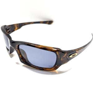 May include: Brown and black sunglasses with a tortoise shell pattern. The sunglasses have a rectangular shape with a gray lens and a gold emblem on the side.
