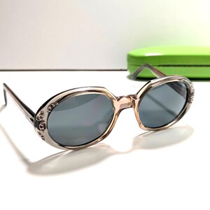 Peut inclure: Lunettes de soleil de forme ovale avec une monture translucide de couleur claire et des verres gris foncé. Les bords extérieurs des montures présentent de petites pierres décoratives incrustées. Un étui rigide vert citron est visible.