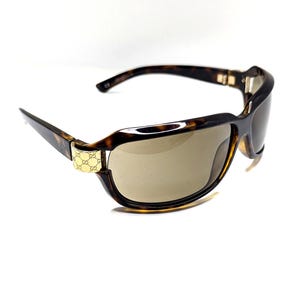 May include: Tortoiseshell sunglasses with brown lenses. The sunglasses have a rectangular frame with a gold-colored accent on the temples. The lenses are a light brown color, and the arms curve slightly.