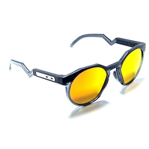 May include: Sunglasses with a dark gray frame and bright yellow-orange reflective lenses. The temples feature a distinct angular, zigzag-shaped design near the hinges, and the Oakley logo is visible on the side of the frame arm.