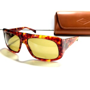May include: Tortoiseshell sunglasses with yellow-tinted lenses. The sunglasses have a rectangular frame and are paired with a brown leather case. The case has a Fossil logo embossed on the front.