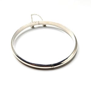May include: A silver bangle bracelet with a simple, elegant design. The bracelet is round and has a clasp closure with a safety chain. The metal has a polished, reflective surface, and the overall style is minimalist and classic.