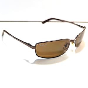 Puede incluir: Gafas de sol rectangulares marrones con lentes tintadas marrones. Las gafas tienen una montura fina de metal marrón oscuro y el logotipo de Ray-Ban es visible en la lente. Las gafas están sobre un fondo blanco.