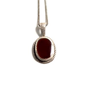 Op de afbeelding: Een zilveren ketting met een ovale hanger met een dieprode steen in een gedraaide metalen rand. De hanger hangt aan een dunne zilveren ketting met vierkante schakels, wat zorgt voor een eenvoudig en klassiek sieraad met een glans.
